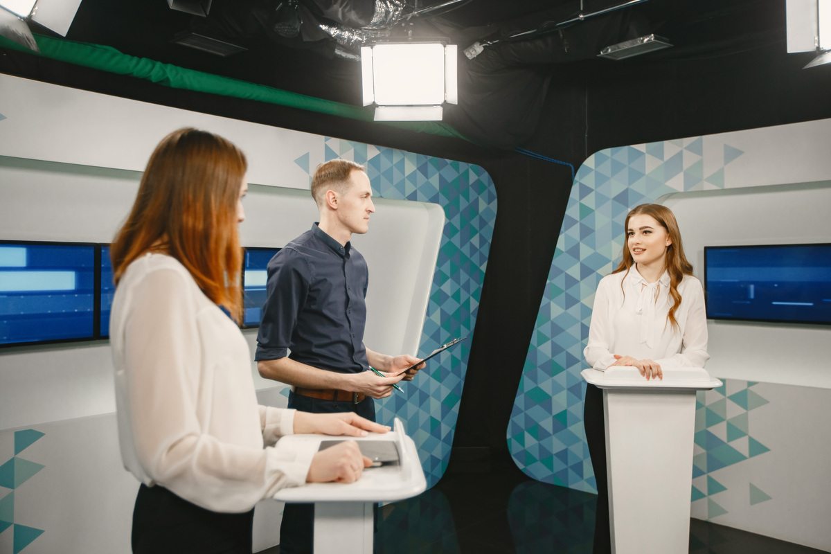 News anchor in studio with two participants answering questions or solving puzzles and host