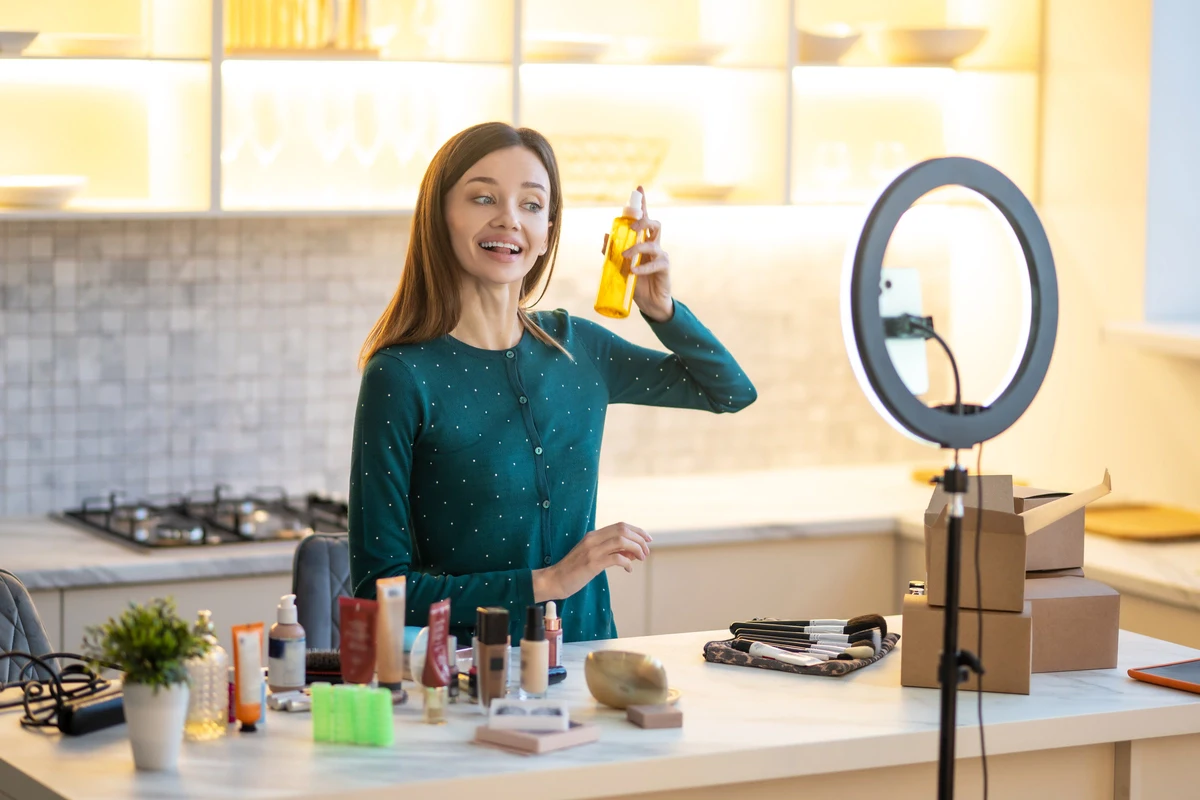 smiling young woman presenting hairspray during online beauty tutorial. Perbedaan global ambassador dan brand ambassador