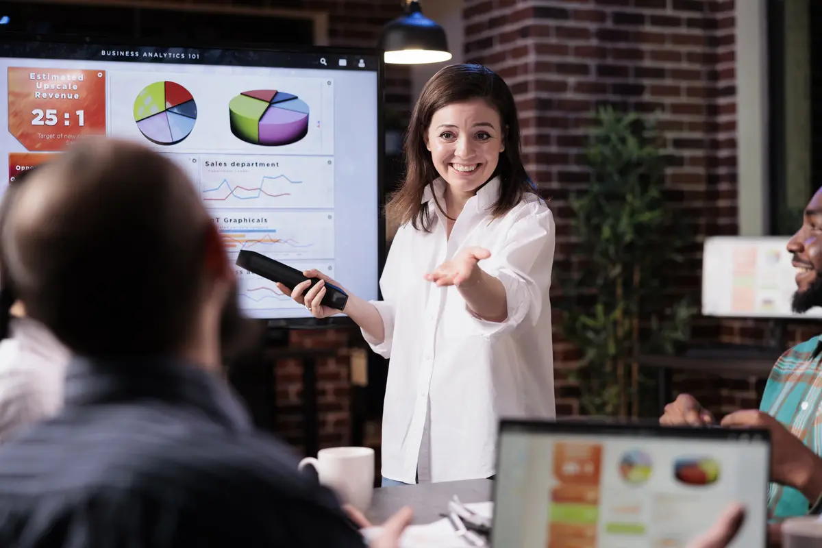 smiling business employee standing in front of tv screen doing elevator pitch with sales statistics talking with man with laptop sitting in late night meeting.
