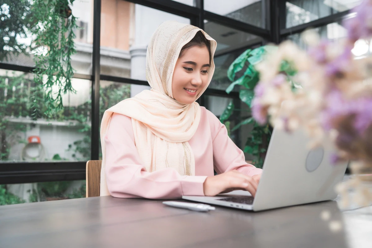 beautiful young smiling asian muslim woman working with laptop sitting in the living room